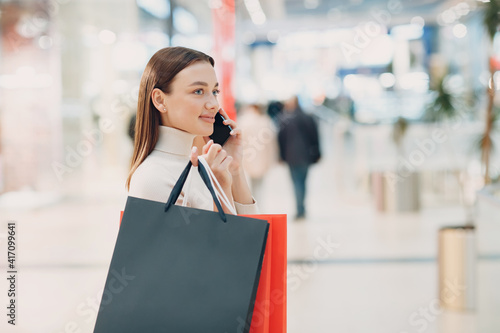 Positive young adult woman carrying paper shopping bags and mobile phone in hands