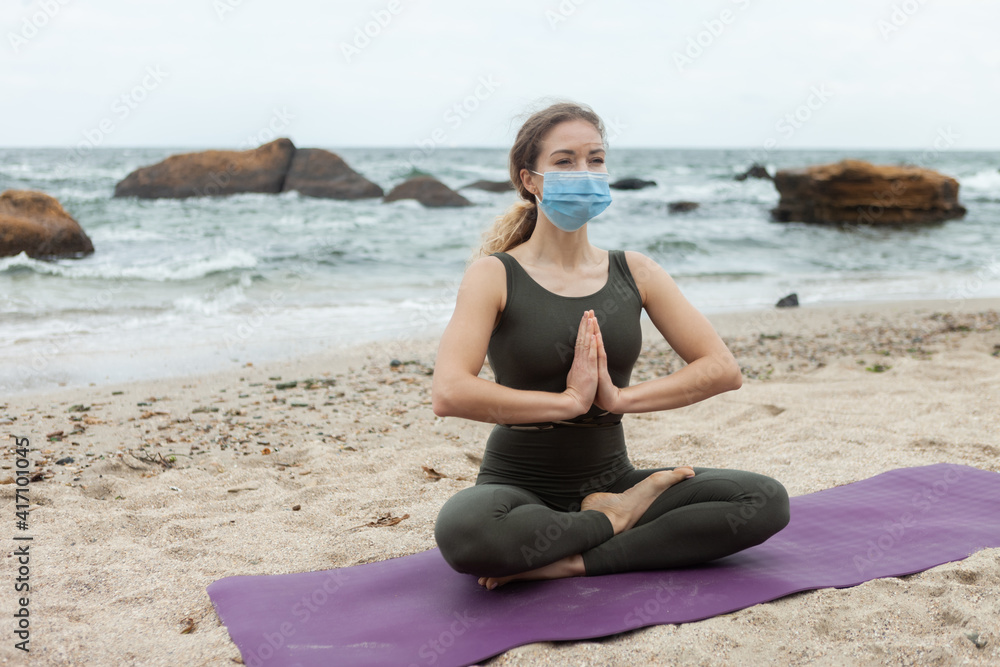 Young yogi woman in sports uniform meditates in a medical mask sitting on mat at beaches. Harmony
