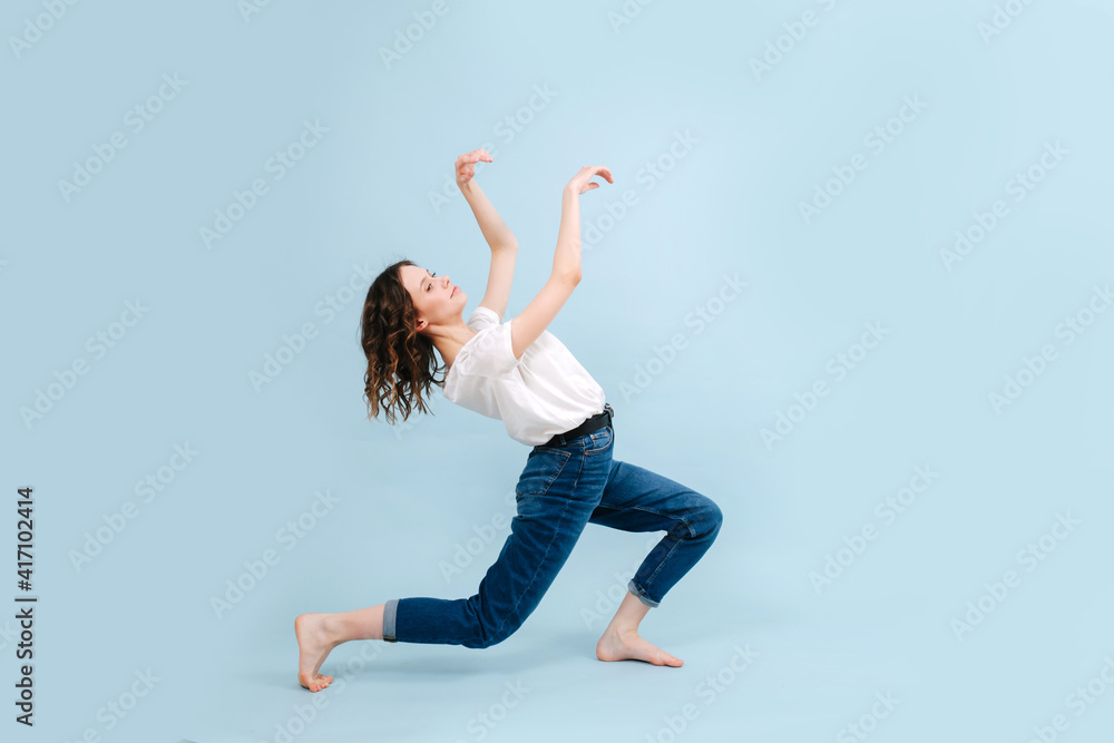 Fototapeta premium Funny contemporary dancer poses in front of blue studio background. Her legs in a lunge, arching herself back, making moves with her hands.