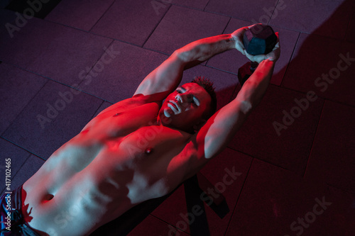 Muscular man exercises with dumbbell lieas on bench in red-blue gradient light in dark cross fit hall photo