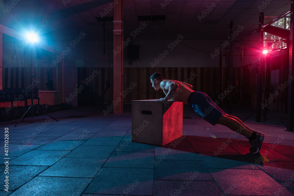 Muscular man training wooden box push-ups in dark gym. Athlete workout in red blue neon light. Healthy lifestyle cross training