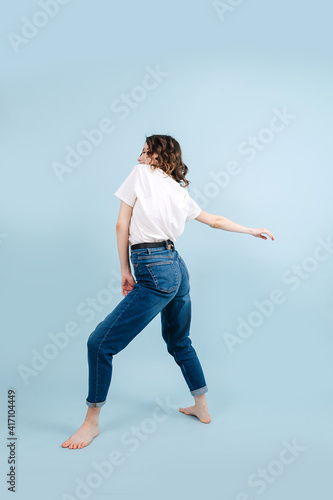 Lively contemporary dancer poses in front of blue studio background. She's lightly arching her back, legs placed solidly, making twisting motion with outstretched hand.