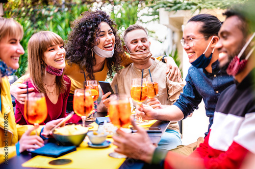 Friends toasting drinks at cocktail bar with face mask - New normal life concept with happy people having fun together drinking spritz at restaurant - Bright end vivid filter - Shallow depth of field