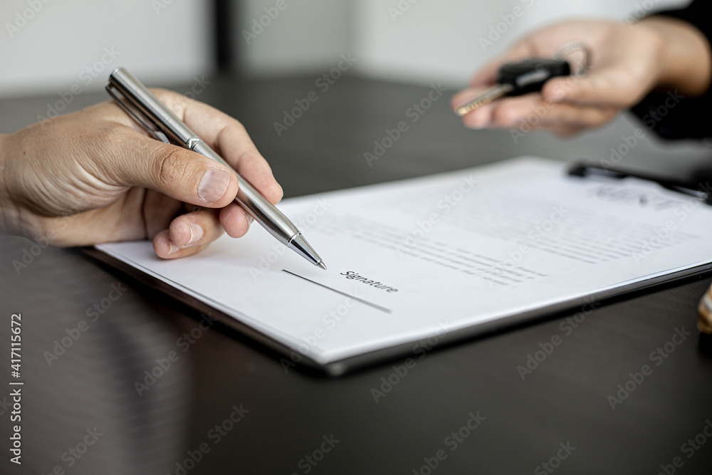 The renter is signing a car rental agreement with the car rental company. After discussing the details and charges with the employee, the employee hand over the car keys to the renter. The renter is signing a car rental agreement with the car rental company. After discussing the details and charges with the employee, the employee hand over the car keys to the renter.
