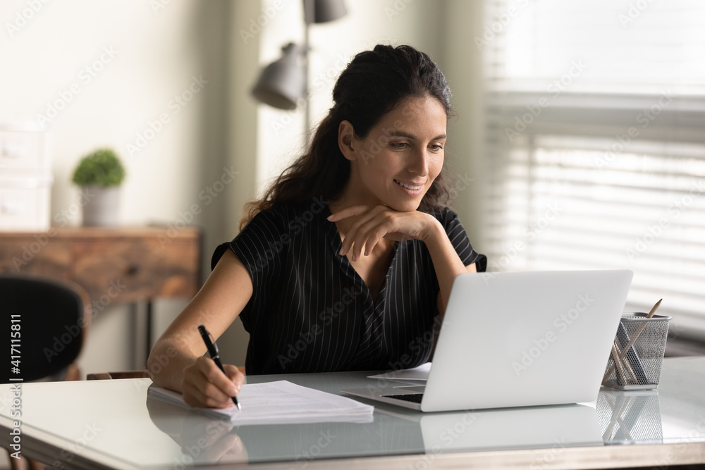 © fizkes - Smiling woman looking at laptop screen, watching webinar or lecture, online course, taking notes, sitting at desk, motivated young female student studying, businesswoman freelancer working on project