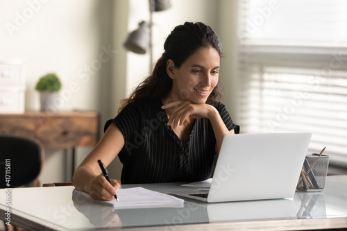 Smiling woman looking at laptop screen, watching webinar or lecture, online course, taking notes, sitting at desk, motivated young female student studying, businesswoman freelancer working on project photo