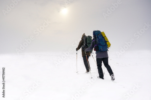 Two guys walk through loose snow during a winter expedition. They carry large backpacks, warm jackets. They hold trekking sticks in their hands.