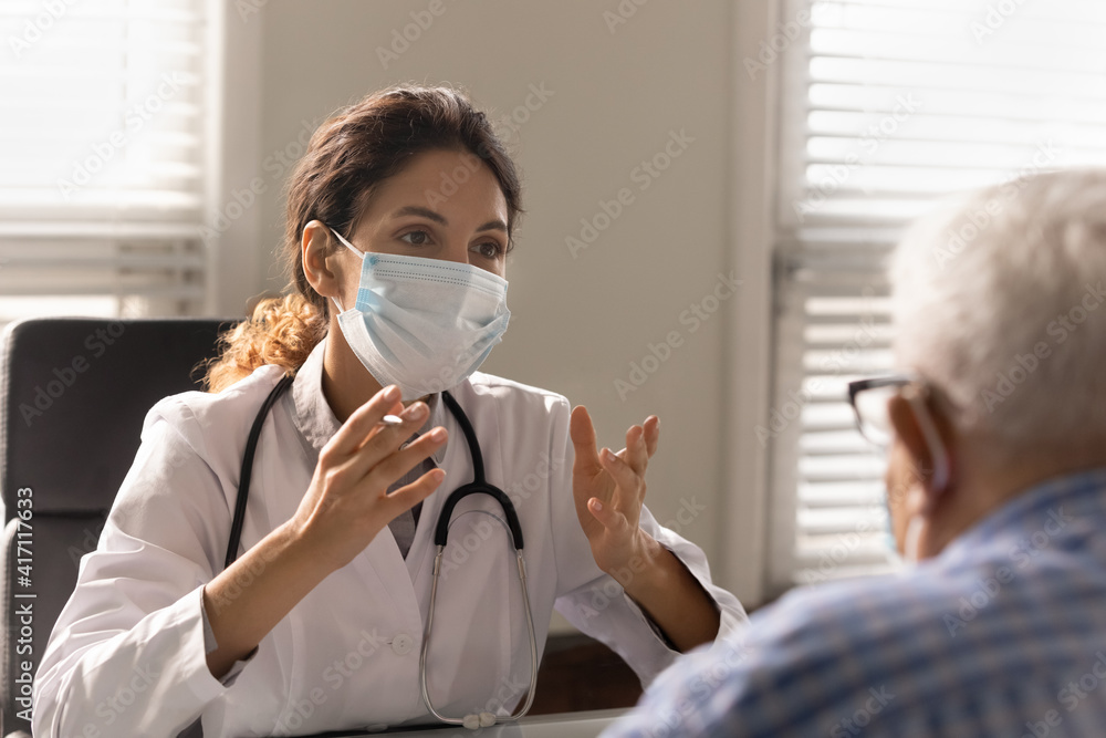 Close up female doctor wearing face mask and uniform consulting elderly patient at meeting in hospital, talking, discussing medical checkup result or symptoms, therapist giving recommendations
