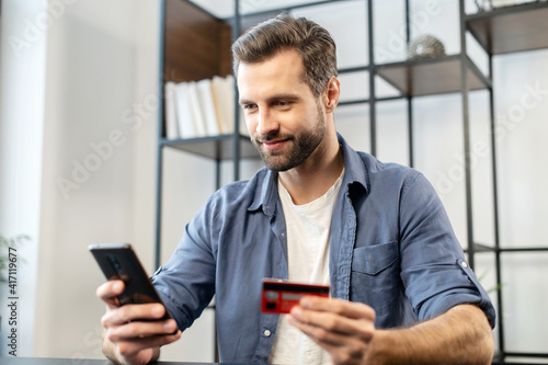 Young bearded hipster guy in casual clothes using mobile phone and credit card for online shopping, entering number to make payment, buying items, holding the debit card for internet banking account