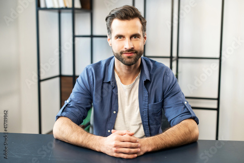 Young bearded guy hipster in casual clothes looking at camera, skilled job applicant is ready for an online interview on a video call, sitting at the desk, holding hands together. Job hunting concept