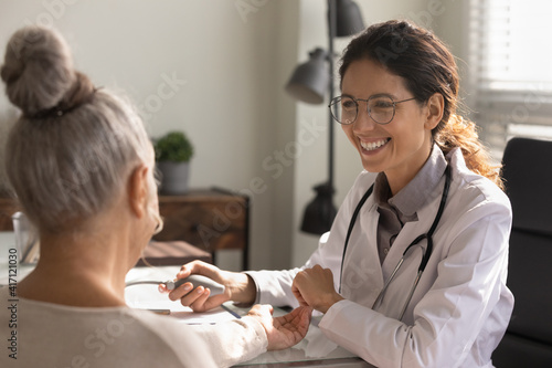Close up smiling female doctor wearing glasses checking old woman blood pressure at meeting in hospital office, using digital tonometer, successful medical checkup results, cardiology healthcare photo