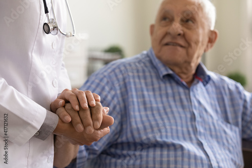 Close up supportive female caregiver wearing white uniform holding mature man hand  giving psychological help  expressing empathy and care  doctor comforting sick senior patient grandfather