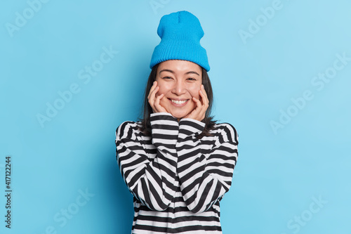 Portait of pleasant looking cheerful Asian woman keeps hands on cheeks smiles gently wears striped jumper stylish hat expresses sincere emotions isolated on blue background. Positive face expressions