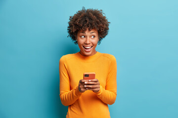 Positive curly haired ethnic woman uses mobile phone checks messages and reads news holds modern cellular in hands looks with curious happy expression on right isolated over blue background.