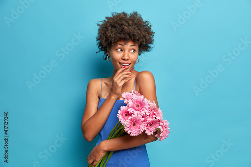 Portrait of lovely dark skinned woman with curly hair wears dress dressed in festive clothes holds bouquet of gerbera flowers on first date looks gladfully aside isolated over blue background