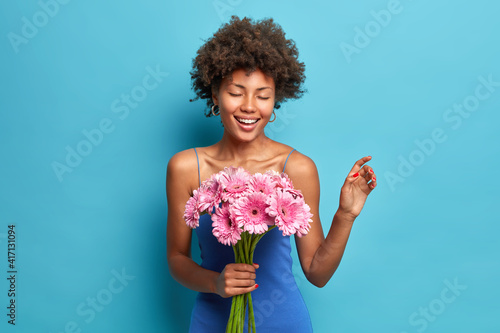 Optimistic beautiful Afro American woman smiles gently holds bouquet of gerberas received as gift on 8 March wears dress closes eyes smiles gently isolated over blue background. Women holidays