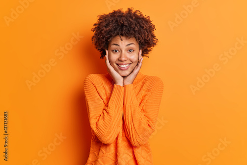 Portrait of pleasant looking dark skinned woman keeps hands on face admires something smiles gently and looks directly at camera wears sweater isolated over vivid orange background. Monochrome © wayhome.studio 