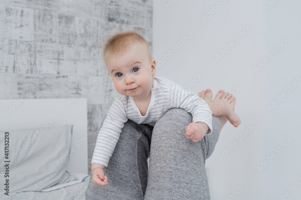 Cute little caucasian baby lying on legs of dad, looking at camera on bed, modern white and gray interior at home. Leisure activities with parent. Copy space. Cute little caucasian baby lying on legs of dad, looking at camera on bed, modern white and gray interior at home. Leisure activities with parent. Copy space.