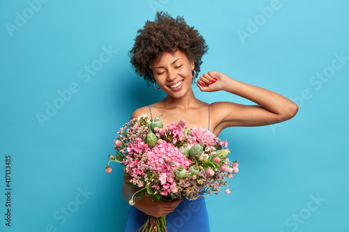 Women and spring time concept. Smiling carefree curly haired Afro American woman celebrates 8 March poses with beautiful bouquet of flowers wears festive dress isolated over blue background.