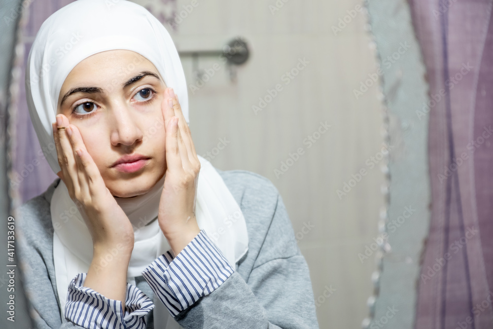 Muslim girl cleaning her face and looking at herself in the mirror Muslim girl cleaning her face and looking at herself in the mirror