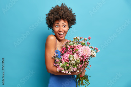 Joyful optimistic beautiful Afro American woman looks happily at camera holds bouquet enjoys spring time dressed in festive clothes isolated over blue background. 8 March concept celebration.