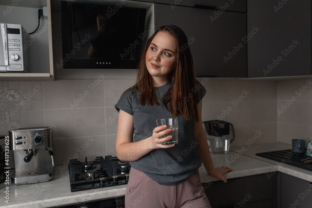 A young brunette woman stands in the kitchen and drinks water from a glass glass A young brunette woman stands in the kitchen and drinks water from a glass glass