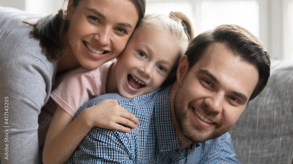 Portrait of cute happy family stacked on couch laughing and looking at camera. Couple of parents and excited kid cuddling, hugging, laughing, having fun at home. Dad piggybacking child and mom.