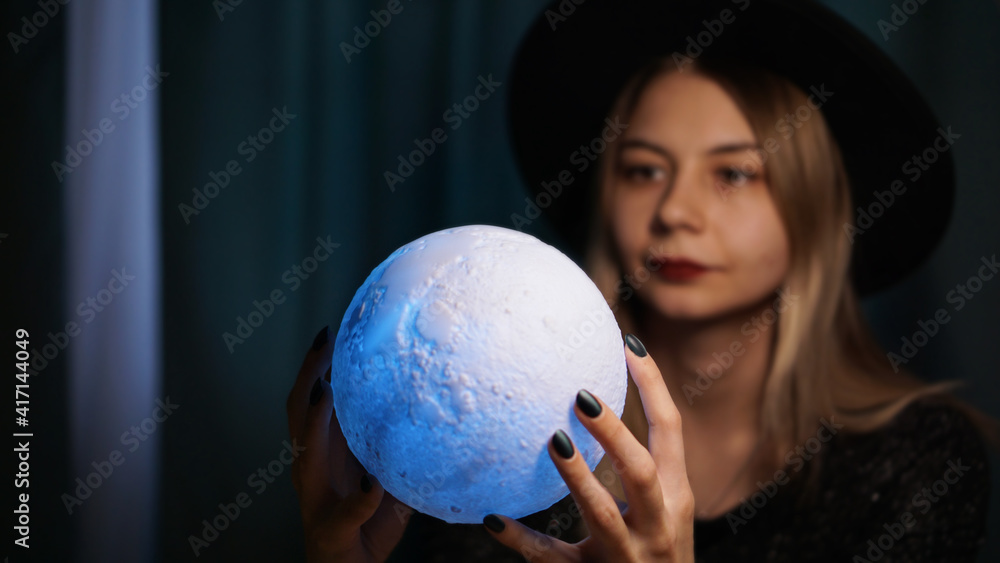Naklejka premium A young woman fortune teller in a hat is holding a magic ball. Beautiful psychic woman is guessing with a blue magic ball