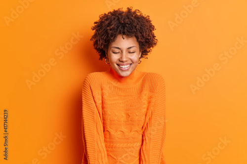 Positive human emotions concept. Cheerful dark skinned beautiful woman closes eyes and smiles broadly wears long sleeved sweater poses against vivid orange background has happy smile on face
