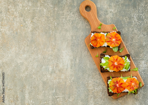 A healthy breakfast or snack, open sandwiches on rye bread with cottage cheese or cream cheese, slices of bloody oranges and herbs on a wooden board on a gray concrete background.