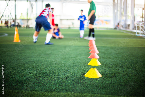 Cone markers is soccer training equipment on green artificial turf with blurry kid players training background. Material for training class of football academy.