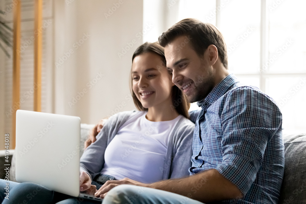 Happy young couple resting and hugging on sofa, watching funny movie online.  Millennial man and woman using laptop for video call from home, shopping on  internet, playing virtual game Stock Photo |