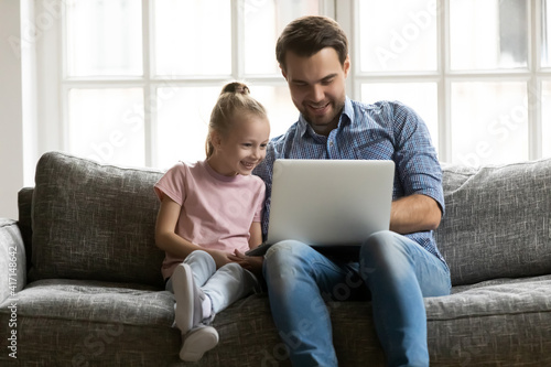 Happy dad and cute preschooler daughter kid sitting on sofa in living room and using laptop for video call. Daddy and girl watching movie on computer, playing online game, shopping on internet