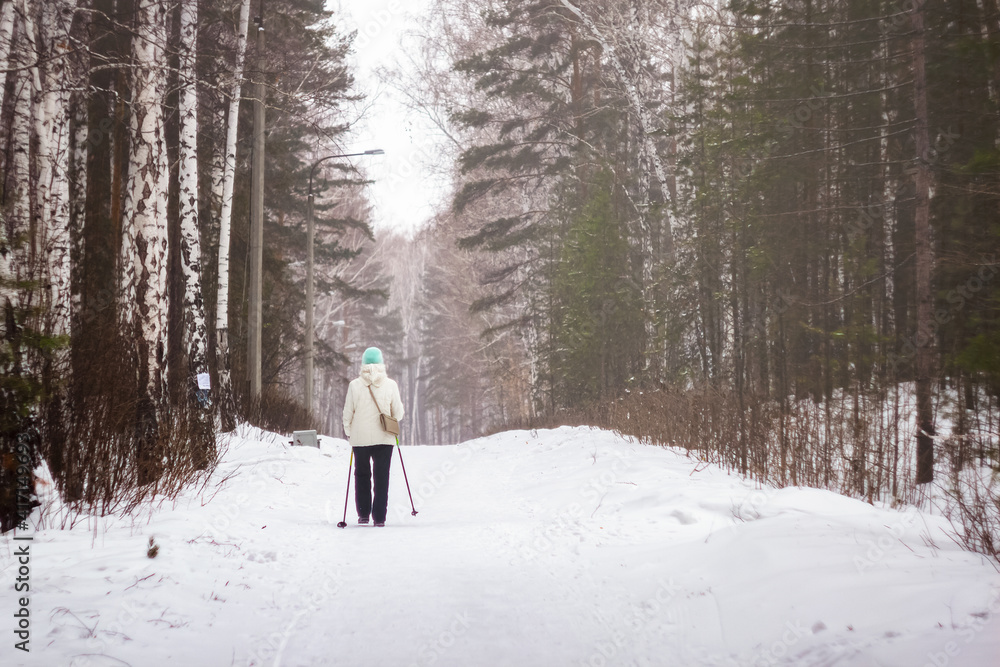 A woman aged in the winter forest is engaged in Scandinavian walking.  active lifestyle in old age.  health promotion.  woman in winter forest.  jogging in the fresh air among the birches.