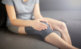 A woman sits on the couch  holds a sick knee with her hands. Problems of orthopedics, disease, trauma