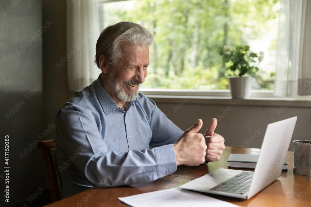 Happy grey haired senior 60s - 70s aged man uses laptop for video call answer from home, shows thumbs up like gesture at webcam, talks, smiles at screen, laughs at web cam, enjoys online virtual chat Happy grey haired senior 60s - 70s aged man uses laptop for video call answer from home, shows thumbs up like gesture at webcam, talks, smiles at screen, laughs at web cam, enjoys online virtual chat