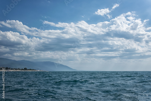 Seascape with waves in the foreground and mountains in the distance. A beautiful blue sky with whimsical white clouds floating in the distance. A small edge of the coast with the beach is visible