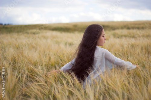 little girl with long dark hairs is walking in sunny summer field - warm landskape with fluffy rye and blue sky