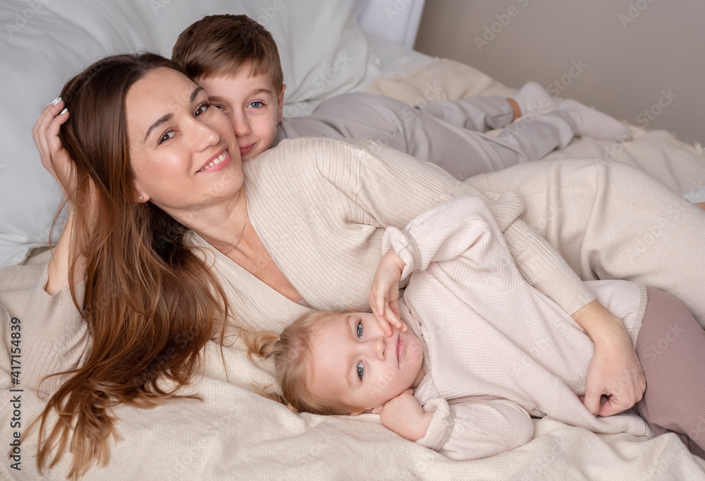 Siblings with their young smiling mother lying on a bed and looking at the camera. Siblings with their young smiling mother lying on a bed and looking at the camera.