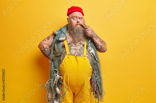 Photos Photo of thoughtful plump seaman poses with fishing net smokes pipe raises eyebrows has pensive expression dressed in overalls tattooed body isolated over yellow background