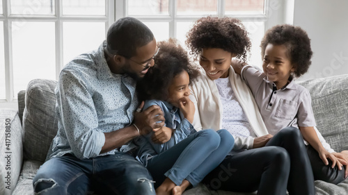 Happy millennial black married couple and two kids enjoying leisure time in living room together. African American parents and children relaxing and hugging on couch at home. Family bonds concept © fizkes