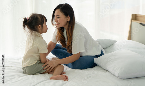 asian mom and young kid with smiling face sitting together on bed in bedroom