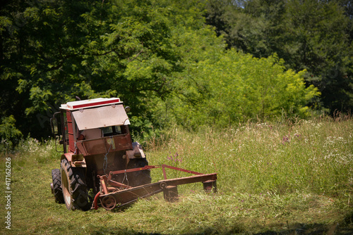 an old red tractor mowing the grass near the forest. use of agricultural machinery on the farm © badescu