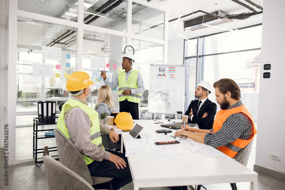 African man in uniform and helmet presenting new construction project to colleagues at office. Three men and one woman sitting at table and listening their competent partner. African man in uniform and helmet presenting new construction project to colleagues at office. Three men and one woman sitting at table and listening their competent partner.