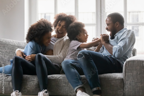 Happy black married couple and kids having fun, tickling on couch in living room. Mom, dad and two kids enjoying leisure at home, sitting on sofa, hugging, cuddling, laughing. Family with children