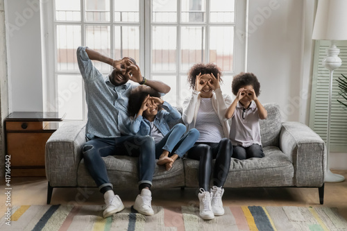 Portrait of happy millennial black couple and kids having fun together at home, making finger glasses. Parents and children sitting on couch, showing binocular eyewear with hands on faces for camera