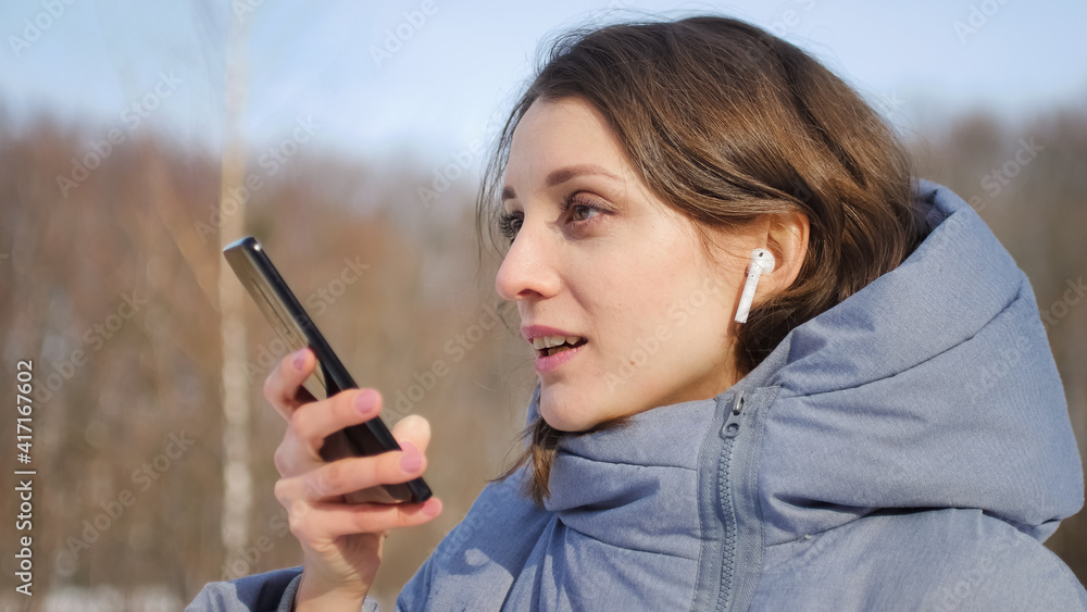 Young woman with wireless headphone is using her smartphone to send a voice message to a new social network Clubhouse standing somewhere outdoors at the park Young woman with wireless headphone is using her smartphone to send a voice message to a new social network Clubhouse standing somewhere outdoors at the park