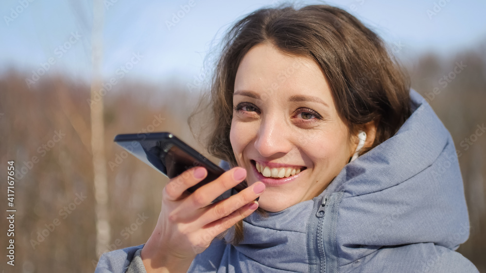 Young woman with wireless headphone is using her smartphone to send a voice message to a new social network Clubhouse standing somewhere outdoors at the park Young woman with wireless headphone is using her smartphone to send a voice message to a new social network Clubhouse standing somewhere outdoors at the park