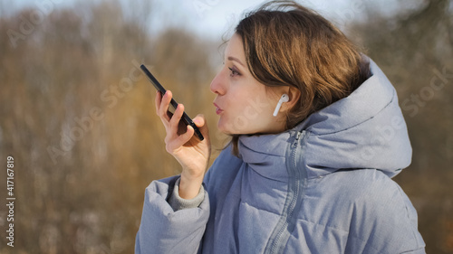 Young woman with wireless headphone is using her smartphone to send a voice message to a new social network Clubhouse standing somewhere outdoors at the park