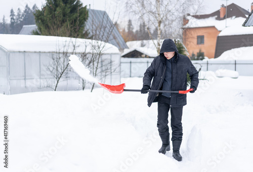 Adult man in black winter clothes shoveling snow in the garden from a sidewalk or driveway with a lightweight red snow shovel, on a cold sunny day.